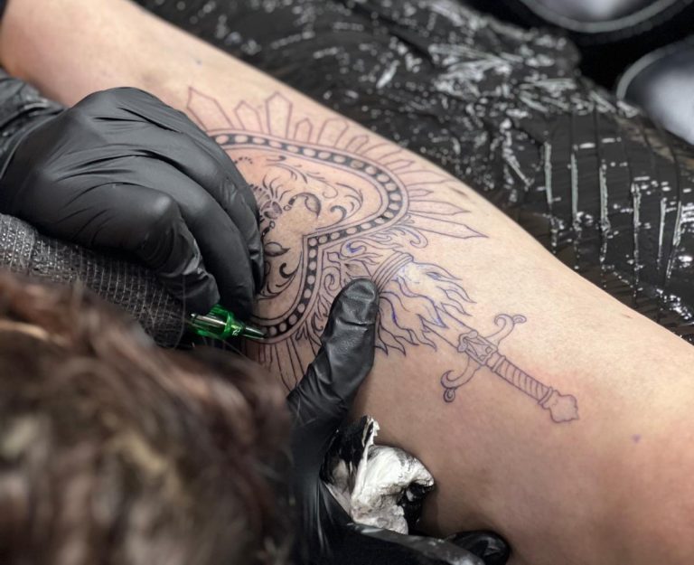 Gros plan d’un tatouage en cours : motif solaire ornemental et épée en linework sur un avant-bras, réalisé avec gants et machine.