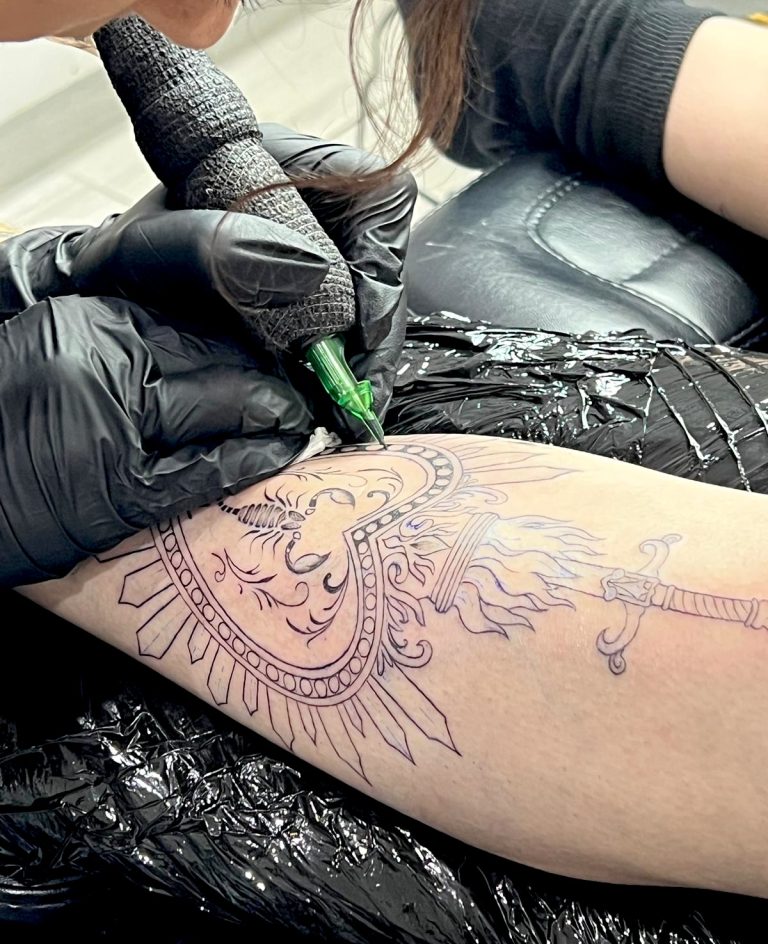 Tatoueuse réalisant un tatouage linework avec un soleil ornemental et une épée sur le mollet
