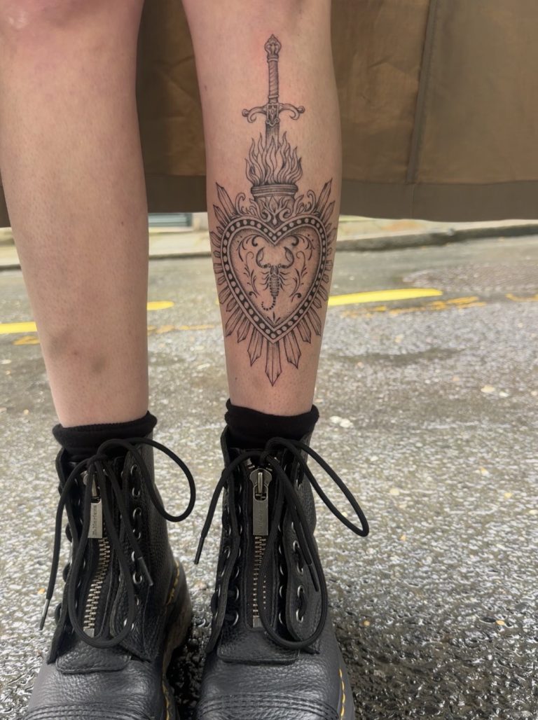Tatouage ornemental noir et gris sur le mollet : cœur rayonnant enflammé, épée au-dessus et scorpion au centre Fougères 35